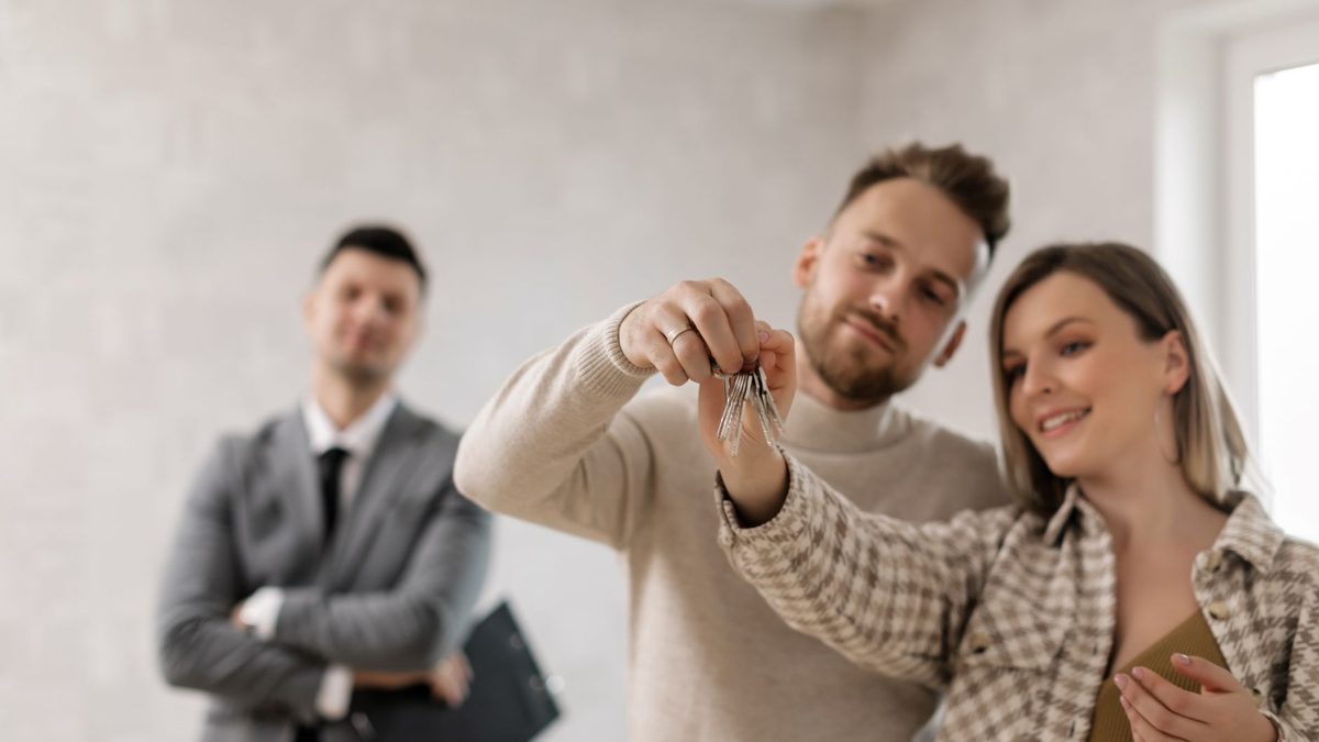 Couple holding keys to their first home with an agent in a bright empty room