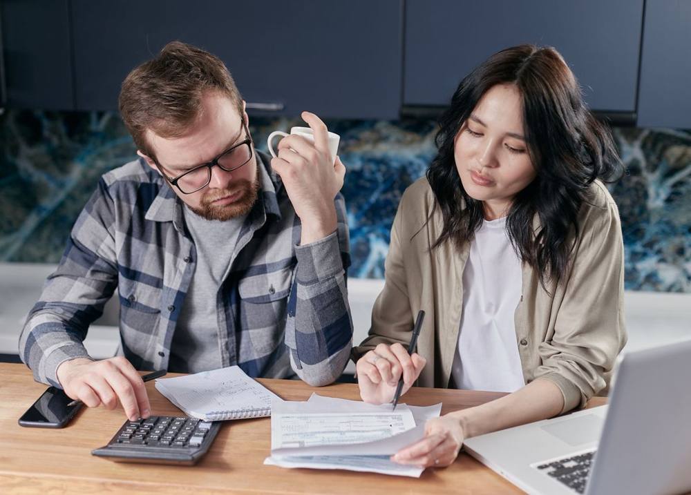 Couple at a kitchen table working through paperwork with a calculator and laptop